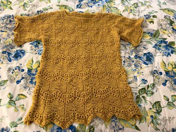 My finished Ripple Tee, a lace top in mustard-gold yarn laid flat on a floral bedspread, showing the scalloped hem and repeating eyelet pattern down the body